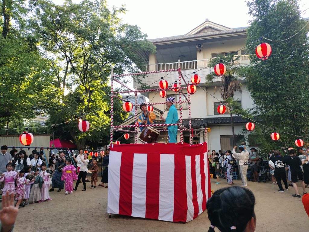 幼稚園敷地内グラウンドにて盆踊り。チンドンかわち家が太鼓を叩き、音頭を取っている